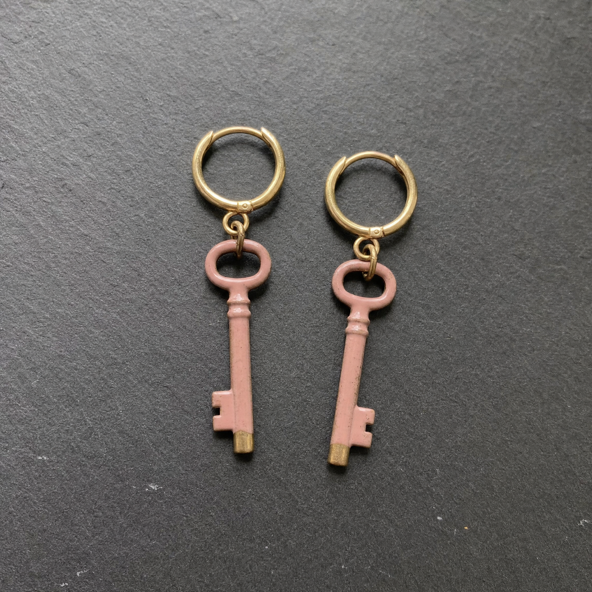 A pair of dusty rose enamelled brass key charm earrings on dark slate