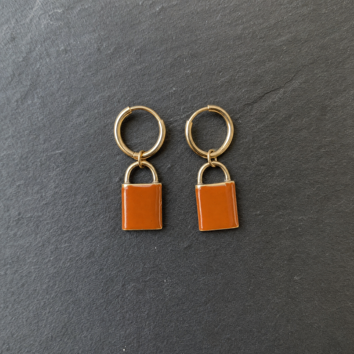 A pair of burnt orange enamelled brass padlock charm earrings on dark slate