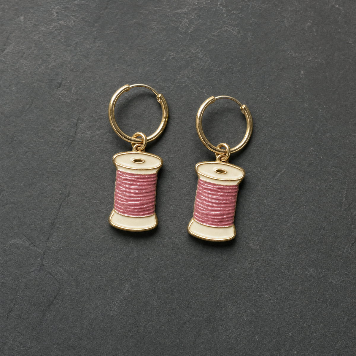 A pair of cream and dusty rose enamelled brass thread spool charm earrings on dark slate
