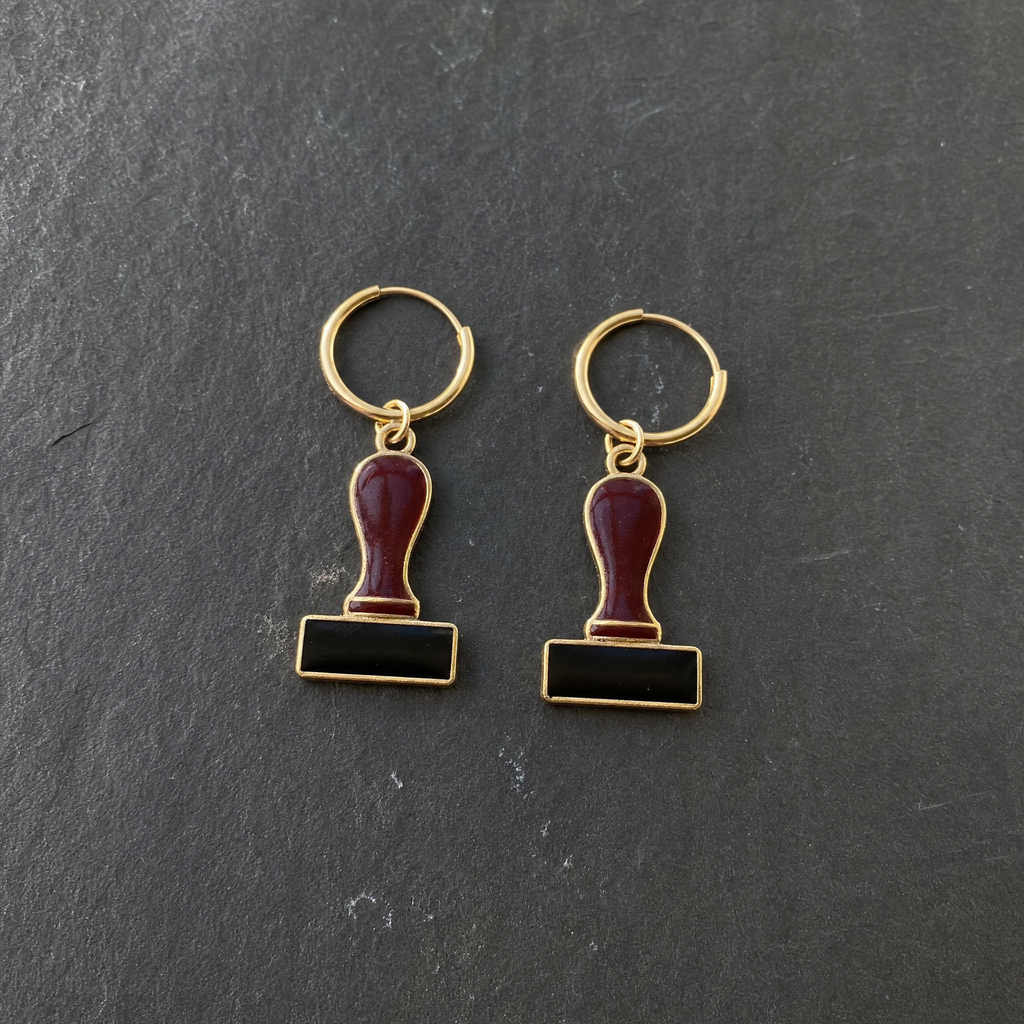 A pair of burgundy and black enamelled brass rubber stamp charm earrings on dark slate