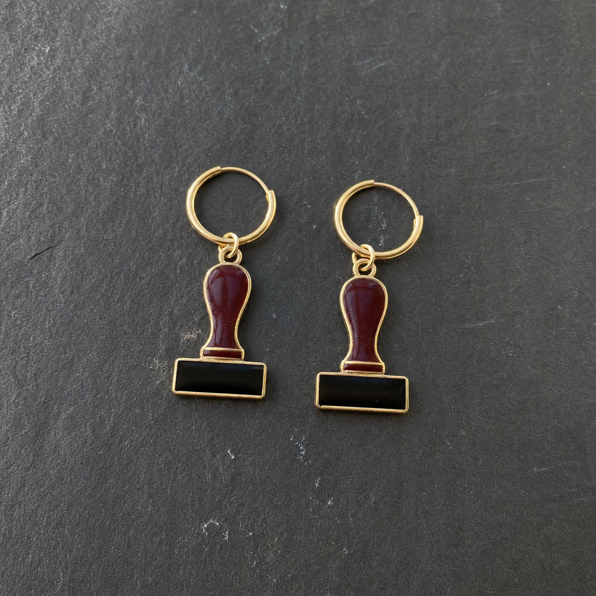 A pair of burgundy and black enamelled brass rubber stamp charm earrings on dark slate